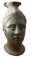 BLS-3003 - Balsamaire en forme de tête fémininebronzeBalsamaire en forme de tête féminine, les traits classiques, les cheveux tirés en arrière et retenus par un bandeau; le col du vase placé au sommet du crâne est équipé de deux bélières coulées diamétralement opposées.