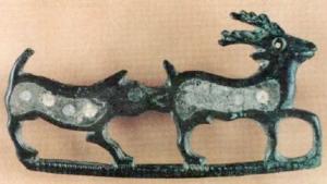 FIB-41457 - Fibule zoomorphe, groupe : chien et cerfbronzeFibule représentant un chien attaquant un cervidé par derrière; barre reliant les pattes des deux animaux, dont les corps sont couverts de loges d'émail incluant des pastilles de verre.