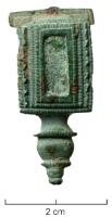 FIB-4221 - Fibule émailléebronzeTPQ : 70 - TAQ : 150Arc en forme de plaque rectangulaire (parfois avec une série de petits ergots sur les côtés), avec une loge d'émail au centre, encadrée de deux lignes ondées ou guillochées; pied en bouton mouluré ou motif trilobé.