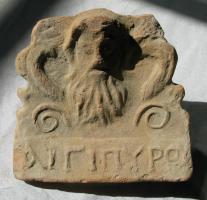 ANT-3002 - Antéfixe : visage barbu, Aigipurouterre cuiteAntéfixe figurant un visage barbu entre crosses et volutes ; en-dessous, inscription moulée AIGIPUROU.