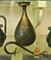 BLS-4017 - BalsamairebronzeTPQ : -25 - TAQ : 25Balsamaire à corps piriforme, col étiré et bord élargi, lèvre en bandeau; anse surélevée, plate et cannelée, rattachée à la panse du vase, en bas, par des appliques en forme de spirales.