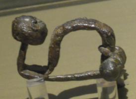 FIB-2031 - Fibule à pied redresséfer, bronzeFibule à pied relevé à angle droit, terminé par un bouton légèrement conique ou hémisphérique; arc à courbure très prononcée; ressort en arbalète entoulé sur une tige en fer présentant une perle en bronze à chaque extrémité.