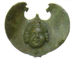 FIB-4279 - Fibule en forme de pelte avec visagebronzeTPQ : 1 - TAQ : 100Broche plate en forme de pelte, lisse, au centre de laquelle est riveté une applique en bas-relief représentant un enfant au visage plein, coiffé d'un corymbe.