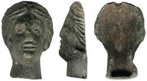 IND-4187 - Applique (?) céphalomorphebronzeApplique en forme de tête féminine, au traits fortement accentués, la chevelure organisée en deux masses séparées par une raie médiane. Au revers, l'objet est creux avec une limite bien définiée à la base du cou; au sommet du crâne, une petite encoche.