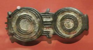 PLB-5594 - Plaque-boucle articulée émailléebronzePlaque-boucle articulée à charnons, constituée de deux motifs circulaires (ornements concentriques), reliés par une plaque.