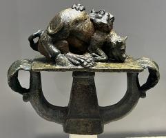 SCH-4030 - Suspension de caisse de char : fauve attaquant un bovidébronzeSuspension de caisse de char constituée d'une scène figurative (lion attaquant un bovidé), posée sur une plaque rectangulaire au-dessus de deux anneaux symétriques terminés par des crosses foliacées ; à la base, douille quadrangulaire.