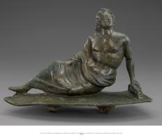 AJG-4051 - Anneau de joug (?)bronzeFigurine (divinité couchée, homme jeune avec un vêtement rabattu sur les hanches et couvrant le bas du corps) posée sur une plaque hexagonale allongée ; au revers, forte bélière hémi-circulaire.
