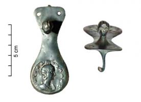 IND-4142 - Objet à dater et à identifierbronzeFibule ? Objet allongé, dont une extrémité arrondie porte un relief monétiforme ; à l'opposé, un buste rapporté entre deux perforation sert à maintenir un porte-ardillon émergeant du revers.