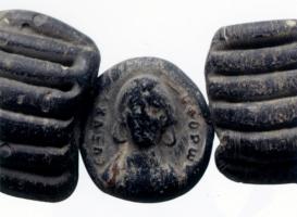 PRL-4017 - Perle monétiformeverrePerle à perforation transversale, en forme de médaillon moulé sur une face : buste féminin de face, à boucles d'oreilles en anneaux, entouré d'une legende en lettres grecques.