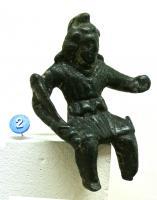 STE-4332 - Statuette : AttisbronzeAttis assis, reconnaissable à son bonnet phrygien et à ssa tunique, tient au creux du bras droit une torche posée sur le genou; le bras gauche est élevé devant lui.