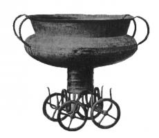 CHV-1004 - Char votifbronzeRécipient de forme variable (chaudron à panse renflée, bord vertical et lèvre déversée ; ou vase à haut pied cylindrique), monté sur un char votif à quatre roues.