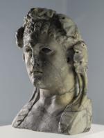 STE-3048 - Buste hermaïque : Héraklès - HerculemarbreLa coiffure comporte plusieurs rangées de petites boucles disposées au-dessus du front. Le jeune homme est imberbe et porte une couronne végétale. La couronne, formée de larges feuilles de vigne réalisées au trépan et ramenées vers l’avant, est retenue par des bandelettes tombant sur les épaules. Le visage s’inscrit dans un ovale régulier et le sillon frontal ainsi que le menton saillant et pointu expriment la virilité du héros. Ce buste hermaïque avec ses mèches courtes, ses traits réguliers et ses yeux en amande semble appartenir aux modèles classiques polyclétéens. Les oreilles sont décollées, dites « en chou fleur », démontrent le caractère combatif du personnage représenté.