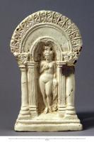 STE-4350 - Vénus dans une niche à sommet arronditerre cuiteLa déesse sort du bain, nue, les cheveux noués au sommet du crâne, une serviette ou un vêtement pendant à la main gauche, la droite posée sur le sein; elle apparaît dans une niche en forme de conque, entre deux pilastres insérés sous une arcade de volutes en S, soutenue par deux colonnes à chapiteaux corinthiens.