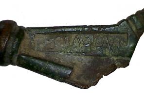 FIB-41239 - Fibule du type Aucissa : HERMOLAVSbronzeTPQ : 1 - TAQ : 100Fibule à arc cintré, bords parallèles, orné de cannelures lisses avec au centre une ligne ondée en relief ; le décor est limité vers le pied par des moulures transversales; porte-ardillon triangulaire plein, marqué d'une estampille, et pied en bouton mouluré rapporté.