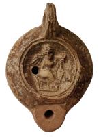 LMP-42677 - Lampe à bec rond limité par un trait : Eros portant une palme, C.ATIL.VESTterre cuiteLampe moulée. Bec arrondi limité par un trait droit ponctué aux extrémités, anse verticale percée à deux sillons sur la tranche. Réservoir circulaire à profil biconvexe. Bandeau penché vers l'extérieur, disque en cuvette limitée par deux sillons concentriques. Base circulaire plate en léger relief limitée par un sillon, estampillée en creux C.ATIL.VEST. Décor : Eros portant une palme.