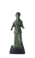 STE-4552 - Statuette : Athéna - Minerve ?bronzeFigurine féminine est en position debout avec pour vêtement un peplos fermé et ceinturé au niveau de la taille. La partie supérieure forme un rabat au-dessus des hanches que l’on nomme apoptygma. Les plis du drapé sont droits et tombants, ils laissent apparaître le genou gauche plié, le personnage se reposant sur sa jambe droite. Le buste possède également un drapé laissant les bras et les avant-bras découverts. La main droite est la moins usée des deux, elle devait probablement présenter un objet. Le haut du corps est très érodé, en particulier le visage dont on distingue difficilement les yeux. Ils sont encadrés par une coiffure dont les mèches sont retenues sur le front et les tempes, puis lâchées en longues boucles sur les épaules et le dos.