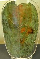 JMB-1002 - JambièrebronzeJambière en tôle, de forme ovale, ornée d'un décor au repoussé comprenant des lignes de ponctuations et des 
