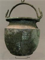 SIT-4048 - Situle de type EhestorfbronzeVase de forme globulaire, presque aussi large que haut, avec un col étranglé et un bord déversé, orné d'une frise moulée ou ciselée; une anse articulée, équipée d'un anneau central, est fixée au col par deux attaches formant une triple feuille vers le bas.