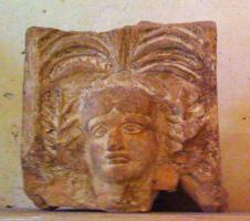 ANT-4024 - Antéfixe : Palmette à 7 branches sur tête
terre cuite
Antéfixe moulée, représentant en façade une tête aux traits fixes (Gorgone ?), surmontée d'une palmette ; palmes de part et d'autre. Parfois, marque épigraphique sous la palmette.