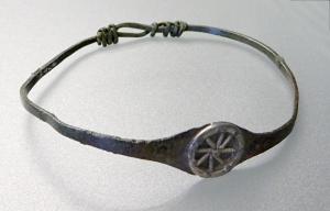 BRC-4118 - Bracelet filiforme à extrémités nouées et médaillon
bronze, argent
TPQ : -30 - TAQ : 200
Bracelet à extrémités nouées, dont le jonc filiforme s'élargit à l'opposé du nœud pour former un médaillon ovale, généralement orné d'un ornement rapporté en argent, de type apotropaïque : roue, croissant..., fixé par un ou deux rivets.