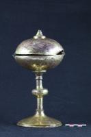 CIB-7001 - Ciboire
cuivre
Ce ciboire est composé d’un pied circulaire à profil concave, d’une haute tige circulaire interrompue par deux petits anneaux encadrant un nœud circulaire, et d’une coupe sphérique à  couvercle à charnière, surmontée d’un tronc de cône supportant une probable croix (disparue) au-dessus d’un petit nœud circulaire.
On retrouve aussi le nœud circulaire à la jonction de la coupe et de la tige. La surface de l’objet est entièrement dorée. La composition du métal reste à analyser, mais tous les exemplaires connus sont en cuivre ou en laiton.