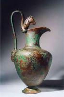 CRU-4018 - Cruche de type Tassinari E.3000
bronze
Aiguière à col étroit et galbé, corps ovoïde à épaule horizontale, posé sur un pied étranglé et mouluré sur sa face inférieure. Le corps et l'anse sont coulés. Le col forme un bec verseur retombant d'un côté. L'anse ornée d'un protomé d'animal (bouc, cheval, griffon...), dont les pattes épousent la forme du col, est surélevée ; son attache inférieure, rattachée sous l'épaule, est en forme de masque (Gorgone ou autre...). La poignée de l'anse peut être décorée de motifs (végétaux...).