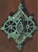 FIB-41088 - Fibule symétrique émaillée
bronze
TPQ : 150 - TAQ : 250
Fibule dont l'arc, de forme générale losangique, résulte d'une composition d'éléments disposés selon un axe de symétrie vertical : au centre, deux roues de char à huit rayons, séparées par un élément losangique émaillé, surélevé; à chaque extrémité, triangles ornés de millefiori; disques émaillées sur tout le pourtour.