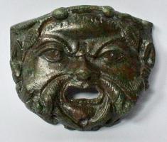 IND-4214 - Déversoir
bronze
Applique légèrement courbe, à sommet rectiligne, figurant une face de Silène (traits grimaçants, couronne de lierre, barbe, oreilles pointues); la bouche largement ouverte sert de déversoir, et sous le menton, trois orifices servent sans doute à augmenter l'écoulement du liquide qui passait par ce déversoir.