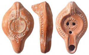 LMP-41670 - Lampe Hayes I : Chrisme
terre cuite
TPQ : 300 - TAQ : 400
Lampe moulée, anse verticale pleine à un sillon, faisant avec la patère une excroissance caractéristique du type. Réservoir piriforme de profil biconvexe. Palmes sur le bandeau. Bec allongé surligné et disque  entourés par un bourrelet, orné d'une couronne striée contenant un chrisme. Base plate limitée par un bourrelet formant une patère avec un double bourrelet rejoignant l'anse. Porte une palmegravée. Motif en S couché entouré de points sous le bec. Engobe rouge brique.