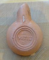 LMP-42306 - Lampe de firme : VETILI
terre cuite
TPQ : 50 - TAQ : 200
Lampe moulée, sans anse. Réservoir circulaire de profil biconvexe asymétrique. Bandeau penché vers l'extérieur porte trois tenons rectangulaires rainurés, non percés, disposés en triangle. Disque en cuvette profonde bordée par un bourrelet qui se poursuite sur le bec le long d'un étroit canal et entoure le méplat. Trou d'évent dans le canal. Base circulaire plate en creux limitée par deux anneaux concentriques enfermant la marque VETILI et parfois un cercle oculé en-dessous.