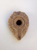 LMP-6010 - Lampe omeyyade miniature à anse recourbéeterre cuiteTPQ : 800 - TAQ : 1000Lampe ovoïde avec bec pointu incorporé dans le réservoir et anse en ruban recourbé, cassée. Épaule convexe. Trou de remplissage défini par un bourrelet circulaire en relief puis par un second, semi-circulaire puis ovale, entourant ainsi aussi bien le trou de remplissage que le trou de mèche. Base plate définie par un anneau en relief en forme de goutte. Ornée de chaque côté de trois demi-cercles entourant des petits disques pointés en relief disposés trois par trois en triangle; entre les demi-cercles, des doubles cercles viennent occuper l'espace libre.
Le canal du bec est décoré de quatre doubles cercles en relief.