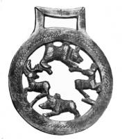 MRS-5001 - Applique latérale de morsbronzeTPQ : 600 - TAQ : 700Applique circiulaire, avec une bélière rectangulaire au sommet pour la fixation des sangles. Le décor ajouré figure une scène de chasse, un chien (?) attaquant un cerf, dans un décor champêtre évoqué par des arbres.