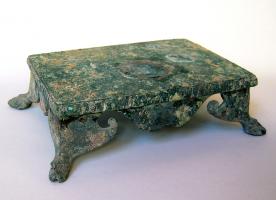 STE-4101 - Statuette : socle
terre cuite
TPQ : 1 - TAQ : 200
Socle quadrangulaire plat, coulé d'un seul tenant, pour statuette de bonne taille (traces de brasures) ; le bord du plateau est godronné; les pieds légèrement obliques ont la forme de sabots de chevaux, prolongés sous les angles par des feuilles à crosse; en façade, crosses découpées.