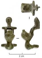 FIB-4285 - Fibule léontomorphebronzeFibule à charnière, arc remplacé par deux protomés de lions adossés, plus ou moins stylisés et posant les pattes antérieures, l'un sur la charnière, l'autre sur une barre transversale; le pied est ici prolongé par une tête humaine très stylisée.