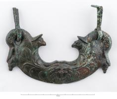 PGM-4018 - Poignée de meuble, tête de MédusebronzePoignée plate à décor figuré, jonc en forme de rameau interrompu par une tête de Méduse (gorgoneion) surmontée de deux têtes de griffons affrontés ; aux extrémités, têtes d'anatidés tenant un gland dans leur bec.