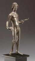 STE-4020 - Statuette : échansonargentFigurine d'un jeune homme nu, en appui sur la jambe gauche et déhanché, tenant un rhyton dans la main droite et une coupe dans la main gauche; lourde coiffure en boucles encadrant le visage.