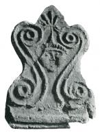 ANT-4003 - Antéfixe : esses et têteterre cuiteAntéfixe moulée, avec en façade deux motifs en S affrontés de part et d'autre d'une tête humaine juvénile ; palmette au-dessus (à la base, parfois, marque du fabricant).