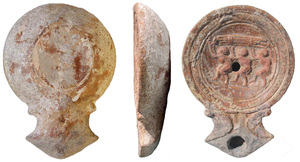 LMP-41759 - Lampe Loeschcke IC : Trois porteurs, amphores terre cuiteTPQ : 50 - TAQ : 100Lampe moulée, sans anse. Réservoir circulaire de profil convexe. Bandeau étroit plat. Disque en profonde cuvette, limité par trois sillons concentrique, décoré de trois porteurs  à gauche portant des amphores protégées par des tiges de bois liées (?) (ou des barres de fer ?). Bec triangulaire à deux volutes simples se poursuivant sous le bec. Base plate circulaire limitée par un sillon. Engobe gris foncé.