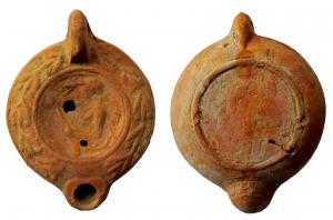 LMP-41984 - Lampe à bec cordiforme : Buste de Satyre, torcheterre cuiteTPQ : 225 - TAQ : 250Lampe moulée, anse verticale pleine. Réservoir circulaire à profil biconvexe., bandeau penché vers l'extérieur au décor de guirlande de laurier noué. Disque en simple cuvette limitée par deux sillons ; buste de Satyre à gauche, tenant une torche.  Bec arrondi cordiforme au dessous strié. Base circulaire plate en léger relief limitée par un bourrelet souligné par deux sillons, et portant un texte incisé sur quatre lignes EX OF[f]I / CINA / ... / ... . Restes d'engobe rouge sur toute la lampe.