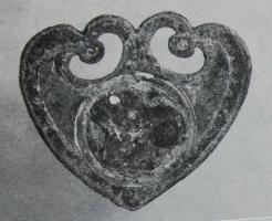 APH-4139 - Applique de harnais : pelteferApplique plate en forme de pelte, les crosses touchant la pointe et donnant aux deux ajours des formes de peltes ; base en ogive; au centre, médaillon circulaire certainement rapporté (tôle estampée ?); au revers, deux pointes de fixation.
