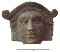 APM-4012 - Applique de meublebronzeApplique en forme de masque de théâtre ou de Lase, aux traits sévères mais figés, la coiffure surmontée d'un ornement terminé par deux crosses sur les côtés ; au revers creux, subsiste l'arrachement d'une barre perpendiculaire.