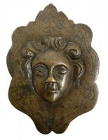 APM-9012 - Applique de meuble : tête juvénilebronzeSur une plaque en forme de fleuron (décor de rinceaux incisés) se détache une tête de jeune garçon, le visage plein, les yeux écarquillés, la coiffure surmontée d'une sorte de feuille dont la pointe émerge au-dessus du front; au revers, barre rectangulaire à extrémité perforée.