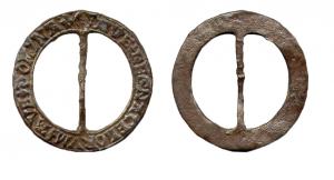 BCC-7001 - Boucle circulaire à traverse, inscritebronzeBoucle circulaire plate, barrée d'une tige mince et fixe; sur la face externe du  pourtour, inscription en lettres gothiques : DOMINA AVE.... Ces exemplaires peuvent être inornés. Elles de distinguent des bouclettes de chaussures par un format plus important, le diamètre dépassant les 30 mm.