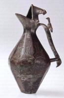 CRU-5015 - Aiguière à panse facettéebronzeCruche coulée, à panse bitronconique, facettée (section décagonale) avec un décor géométrique incisé, dont le bec étiré est protégé par un couvercle partiel, fixe; anse rubanée en forme d'étrier, avec une figurine d'oiseau en guise de poucier.