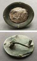 FIB-41810 - Fibule à médaillon : Louve et Jumeauxbronze, argentFibule à charnière entre deux plaquettes coulées, abritant un médaillon ovale maintenu par un cadre plat fixé par quatre rivets : tôle emboutie : la Louve allaitant Romulus et Remus. 