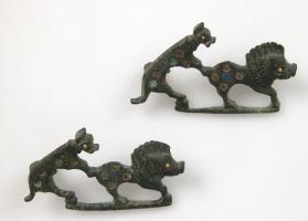 FIB-4293 - Fibule zoomorphe, groupe : chien et sanglierbronzeFibule représentant un chien attaquant un sanglier par derrière, les griffes plantées sur l'arrière train; barre reliant les pattes des deux animaux, dont les corps sont couverts de pastilles d'émail.