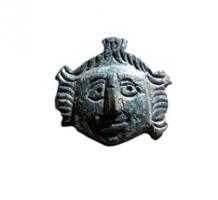 FIB-4580 - Fibule anthropomorphebronzeBroche en forme de visage d'enfant (putto ?), large et encadré de chevelures formant des toupets sur les tempes, avec un corymbe au-dessus du front; détails marqués par de profondes incisions.