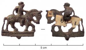 FIB-4685 - Fibule zoomorphe, groupe : cavalierbronzeFibule en forme de cheval monté par un cavalier; le style, assez réaliste, évoque un voyageur marchant au pas; le cavalier bien proportionné tient les rênes, derrière lui une cape forme un arc de cercle rattaché à l'arrière-train de sa monture; la queue et les quatre pattes sont reliées par une barre rectiligne moulurée. Porte-ardillon en plaque creusée d'une fente oblique.