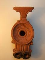 LMP-3131 - Lampe à deux becsterre cuiteLampe à deux becs ronds accostés; disque orné d'une couronne de palmes incisée. Trou de remplissage défini par un haut bourrelet circulaire en relief., épaule convexe. A l'arrière, longue anse verticale moulée, striée en forme de T. Argile brique avec mica, engobe orange foncé; base plate entourée par un anneau en relief. Anse avec réflecteur en forme de T.