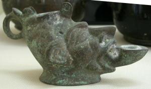 LMP-4021 - Lampe anthropomorphe : tête de FaunebronzeLampe en bronze en forme de tête faunique, aux traits accentués; le bec émerge de la bouche aux lèvres épaisses; couvercle articulé au sommet du crâne; anse en anneau.